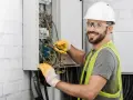 Uśmiechnięty elektryk w kasku i kamizelce pracuje nad instalacją elektryczną w domu, dbając o bezpieczeństwo i porządek.