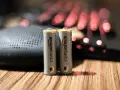 Dwie baterie paluszki AA AmazonBasics stoją na drewnianym blacie, obok głośnika i podświetlanej klawiatury.