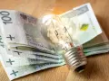 Żarówka na stosie banknotów 100 zł. Symbolizuje wysokie koszty energii i potrzebę wsparcia, np. dodatek energetyczny.