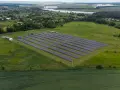 Rozległa farma fotowoltaiczna na zielonym polu, z rzędami paneli słonecznych. W tle widać jezioro i zabudowania.