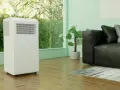 Biały klimatyzator przenośny stoi obok czarnej kanapy. Czy warto zainwestować w taki sprzęt?