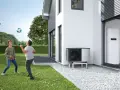 Dwaj chłopcy grają w piłkę na trawniku przed nowoczesnym domem. Obok budynku stoi pompa ciepła, która może obniżyć koszt ogrzewania domu 100m2.
