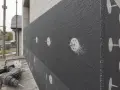 Na budowie trwa kołkowanie styropianu. Widać szarą płytę izolacyjną przymocowaną do ściany białymi kołkami. Obok leży wiertarko-wkrętarka.