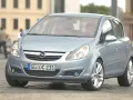 Jasnoniebieski Opel Corsa D z włączonymi żarówkami z przodu, zaparkowany na bruku.