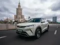 Biały, nowoczesny chiński samochód elektryczny BYD jedzie ulicą w Warszawie, z Pałacem Kultury w tle.
