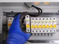Ręka w niebieskiej rękawicy montuje bezpiecznik w skrzynce elektrycznej. Obok widać rząd innych bezpieczników i przewody. To ważny element instalacji, chroniący przed skutkami wyładowań, jak odgromnik.