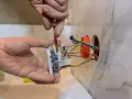 Elektryk montuje białe gniazdko, przykręcając je śrubokrętem. Widać kolorowe przewody wychodzące z puszki instalacyjnej.