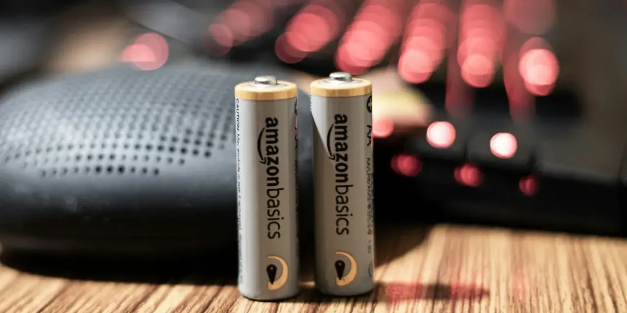 Dwie baterie paluszki AA AmazonBasics stoją na drewnianym blacie, obok głośnika i podświetlanej klawiatury.