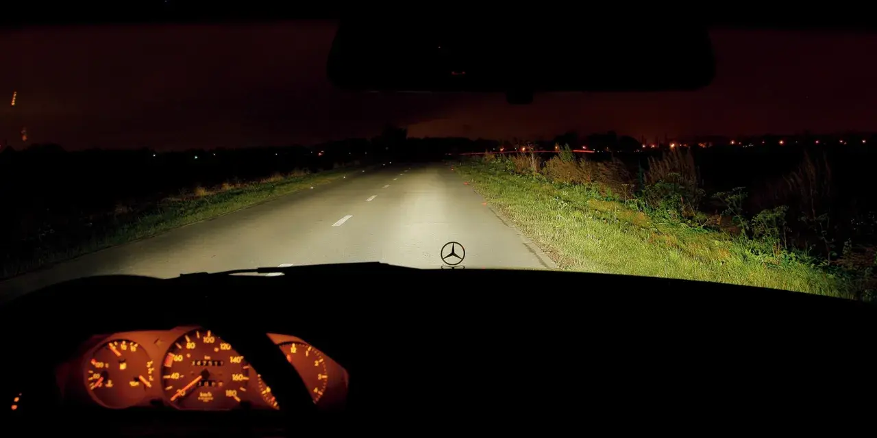 Widok z wnętrza samochodu nocą. Włączone światła drogowe oświetlają pustą drogę. Na desce rozdzielczej widać podświetlone zegary.
