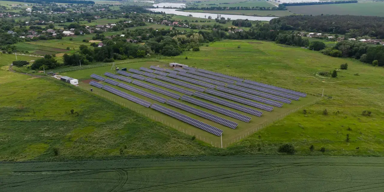 Rozległa farma fotowoltaiczna na zielonym polu, z rzędami paneli słonecznych. W tle widać jezioro i zabudowania.