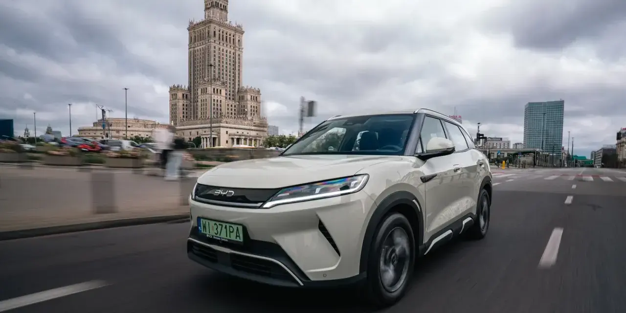 Biały, nowoczesny chiński samochód elektryczny BYD jedzie ulicą w Warszawie, z Pałacem Kultury w tle.