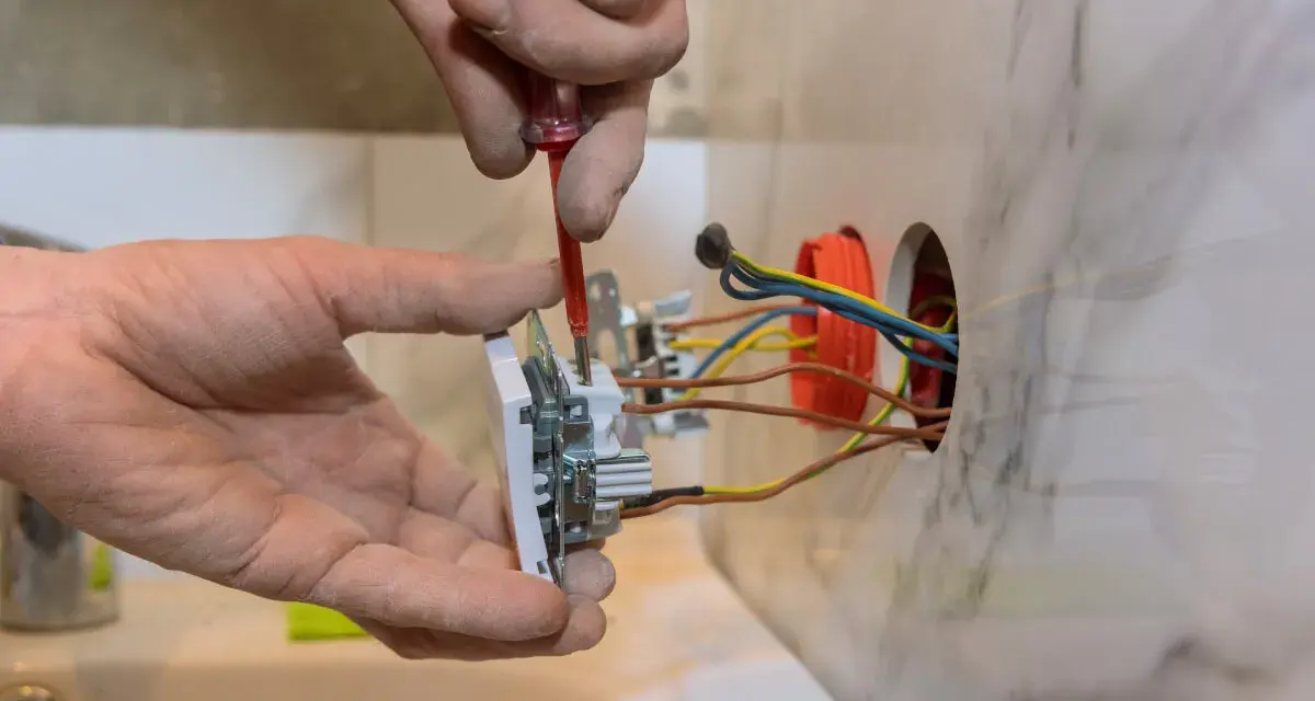 Elektryk montuje białe gniazdko, przykręcając je śrubokrętem. Widać kolorowe przewody wychodzące z puszki instalacyjnej.