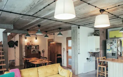 Nowoczesne wnętrze z widoczną instalacją elektryczną natynkową na betonowym suficie. Wygodna kanapa, stół jadalniany i kuchnia.