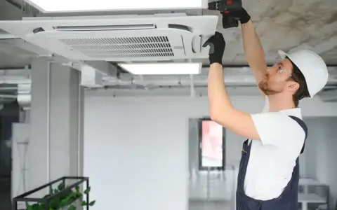 Technik w kasku montuje klimatyzator kasetonowy na suficie.