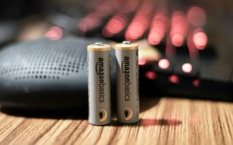 Dwie baterie paluszki AA AmazonBasics stoją na drewnianym blacie, obok głośnika i podświetlanej klawiatury.