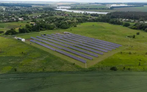 Rozległa farma fotowoltaiczna na zielonym polu, z rzędami paneli słonecznych. W tle widać jezioro i zabudowania.