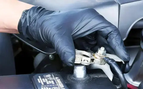 Ręka w czarnej rękawiczce dokręca śrubę na klemie akumulatora samochodowego.