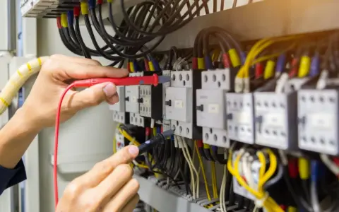 Elektryk sprawdza przewód neutralny w szafie sterowniczej, używając multimetru. Widać wiele kabli i przekaźników.