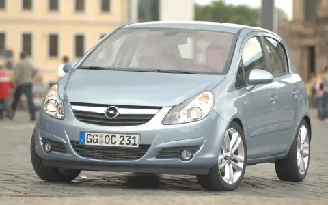 Jasnoniebieski Opel Corsa D z włączonymi żarówkami z przodu, zaparkowany na bruku.