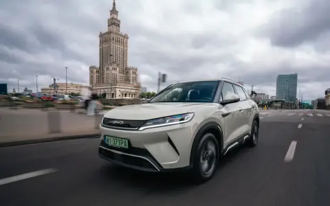 Biały, nowoczesny chiński samochód elektryczny BYD jedzie ulicą w Warszawie, z Pałacem Kultury w tle.