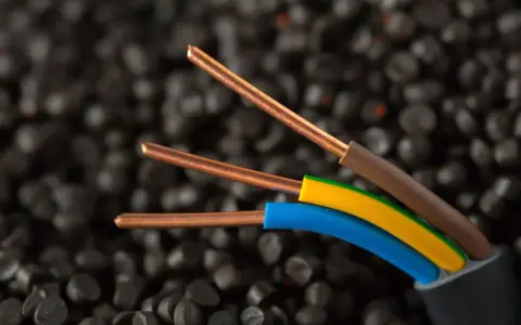 Trzy miedziane żyły kabla elektrycznego, w izolacjach brązowej, żółto-zielonej i niebieskiej, na tle granulatów tworzywa sztucznego. Idealne do instalacji IP20.