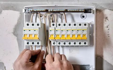 Ręce elektryka podłączają przewody do skrzynki z bezpiecznikami. Widać wlz, czyli rozdzielnicę z wyłącznikami nadprądowymi.