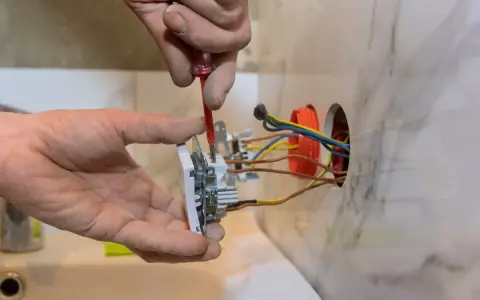 Elektryk montuje białe gniazdko, przykręcając je śrubokrętem. Widać kolorowe przewody wychodzące z puszki instalacyjnej.