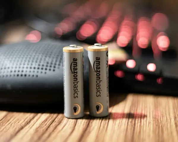 Dwie baterie paluszki AA AmazonBasics stoją na drewnianym blacie, obok głośnika i podświetlanej klawiatury.