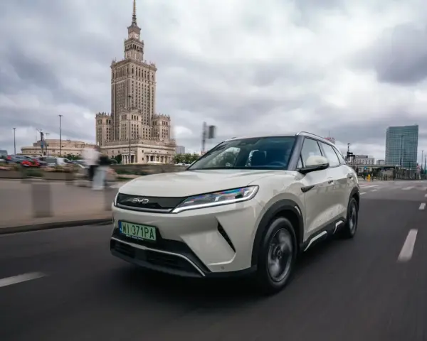 Biały, nowoczesny chiński samochód elektryczny BYD jedzie ulicą w Warszawie, z Pałacem Kultury w tle.