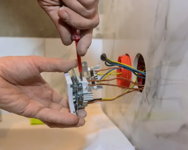 Elektryk montuje białe gniazdko, przykręcając je śrubokrętem. Widać kolorowe przewody wychodzące z puszki instalacyjnej.