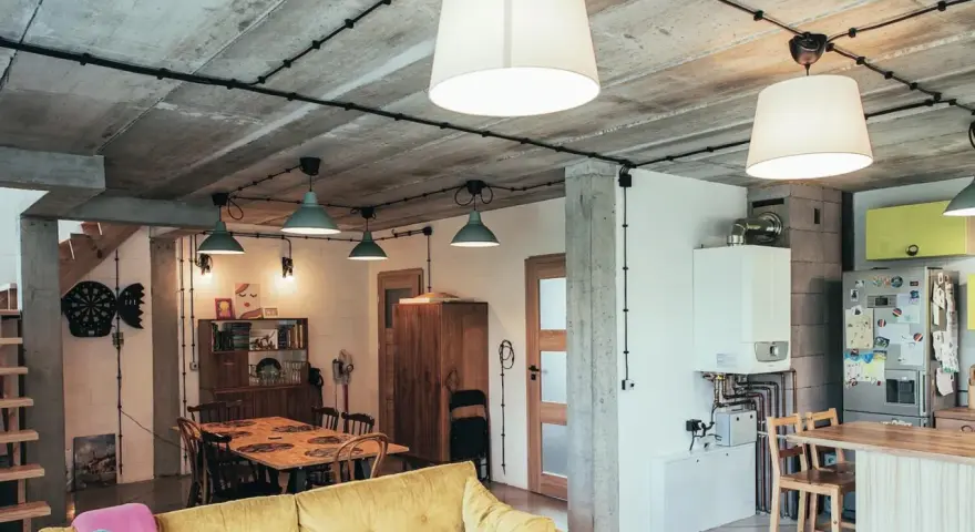 Nowoczesne wnętrze z widoczną instalacją elektryczną natynkową na betonowym suficie. Wygodna kanapa, stół jadalniany i kuchnia.