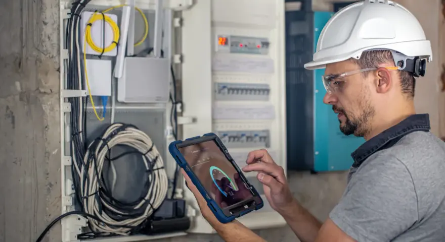 Technik w kasku i okularach ochronnych sprawdza przegląd instalacji elektrycznej domu na tablecie. Widoczne przewody i rozdzielnica.