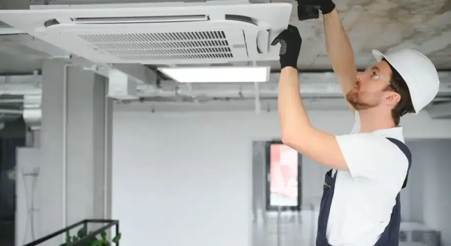 Technik w kasku montuje klimatyzator kasetonowy na suficie.