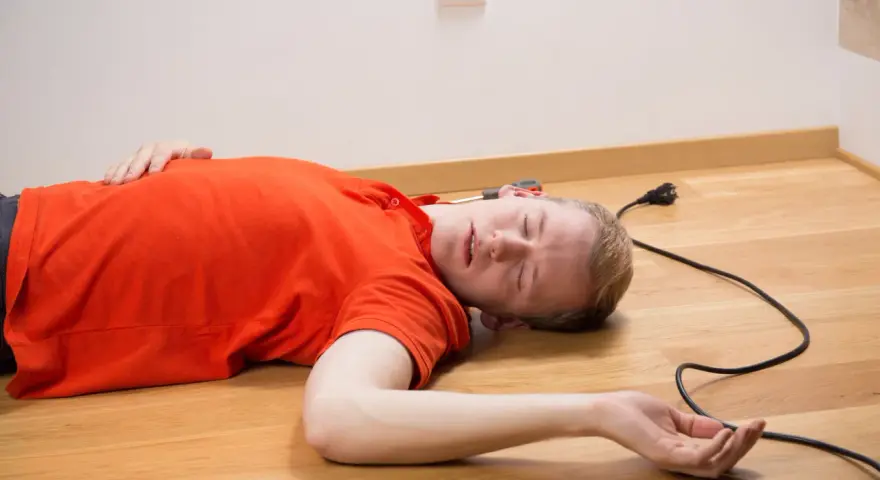 Mężczyzna w pomarańczowej koszulce leży na podłodze, obok kabel z wtyczką. Wygląda na to, że doświadczył porażenie prądem.