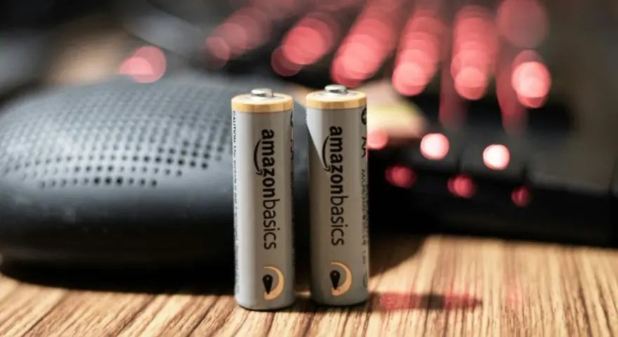 Dwie baterie paluszki AA AmazonBasics stoją na drewnianym blacie, obok głośnika i podświetlanej klawiatury.