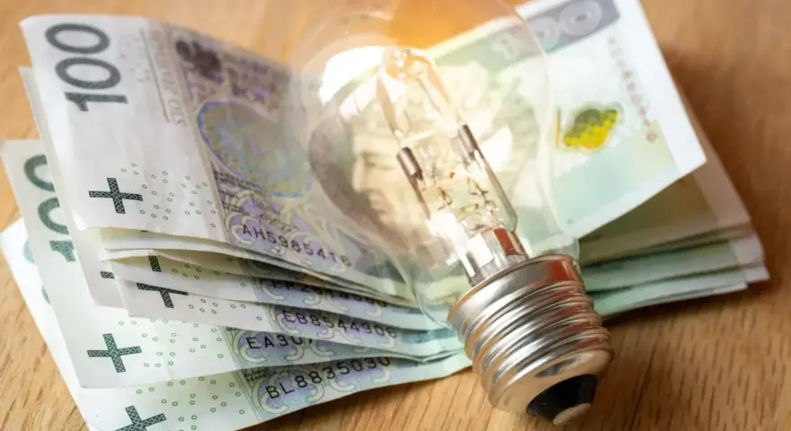 Żarówka na stosie banknotów 100 zł. Symbolizuje wysokie koszty energii i potrzebę wsparcia, np. dodatek energetyczny.