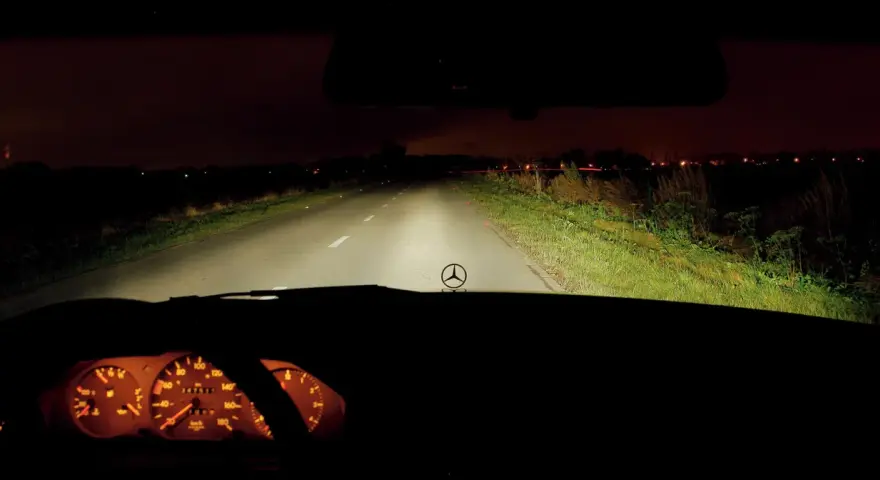 Widok z wnętrza samochodu nocą. Włączone światła drogowe oświetlają pustą drogę. Na desce rozdzielczej widać podświetlone zegary.