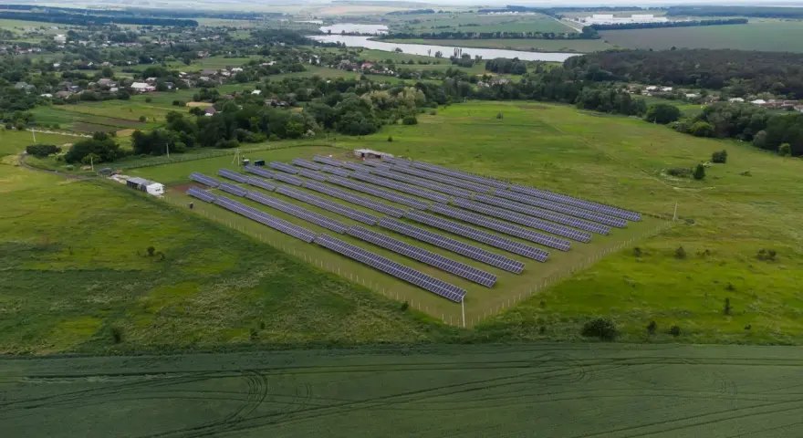 Rozległa farma fotowoltaiczna na zielonym polu, z rzędami paneli słonecznych. W tle widać jezioro i zabudowania.
