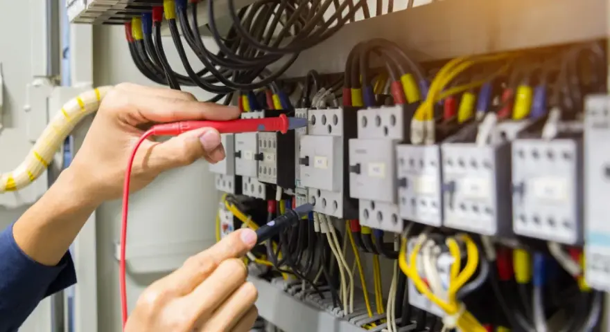 Elektryk sprawdza przewód neutralny w szafie sterowniczej, używając multimetru. Widać wiele kabli i przekaźników.