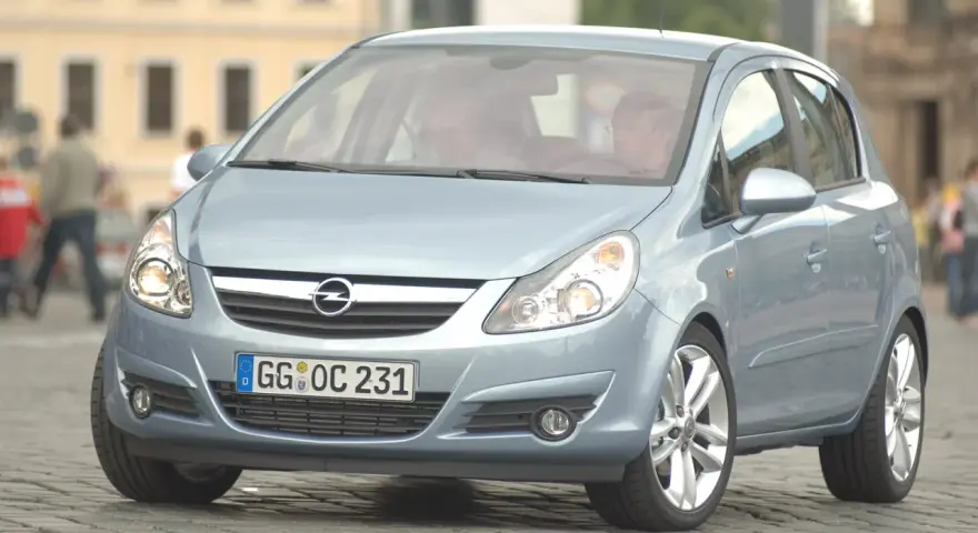 Jasnoniebieski Opel Corsa D z włączonymi żarówkami z przodu, zaparkowany na bruku.