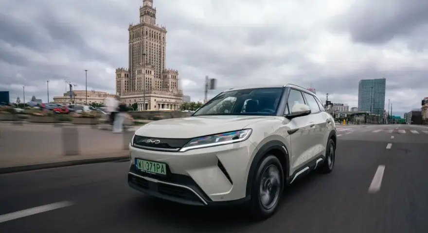 Biały, nowoczesny chiński samochód elektryczny BYD jedzie ulicą w Warszawie, z Pałacem Kultury w tle.