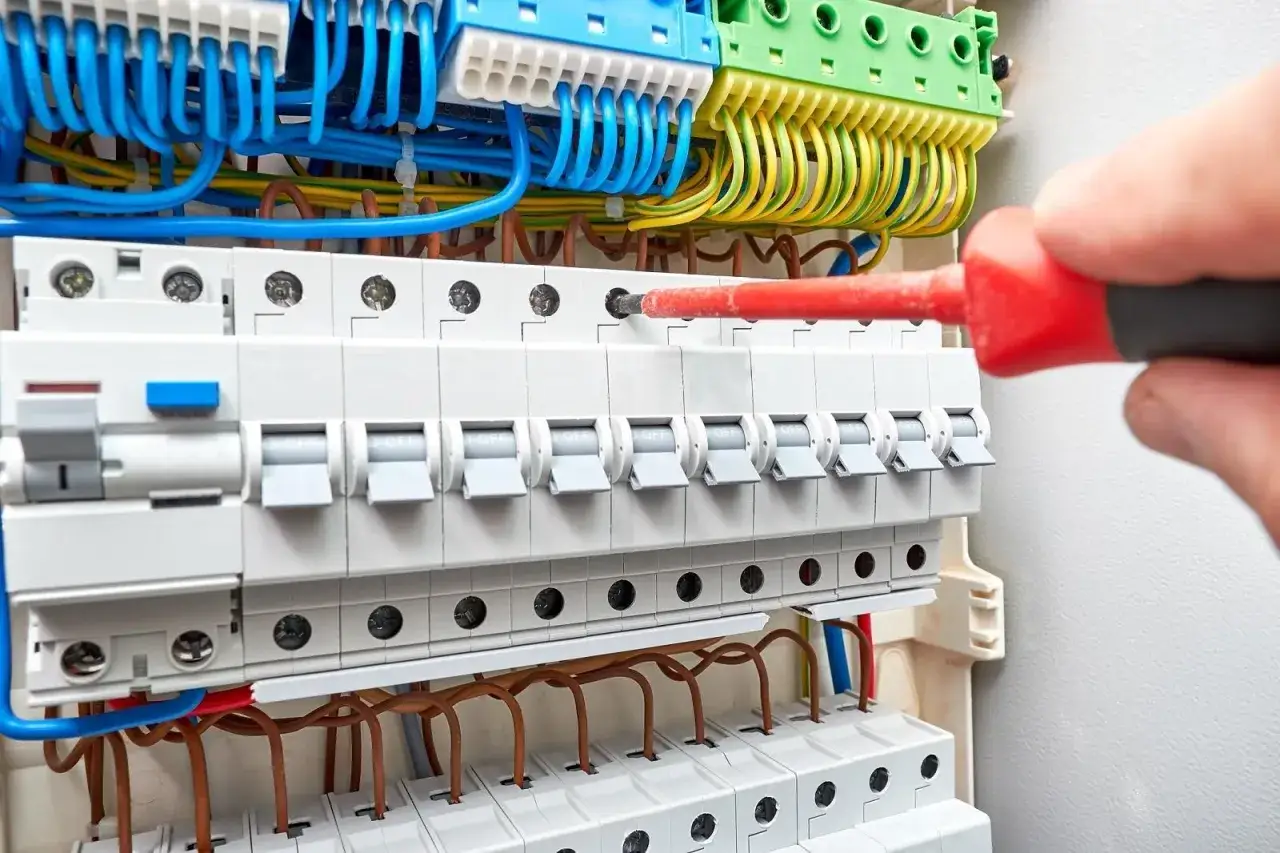 Elektryk dokręca śrubę w skrzynce z bezpiecznikami, gdzie widać przewody i wyłączniki. Instalacja elektryczna IP20 jest gotowa do pracy.