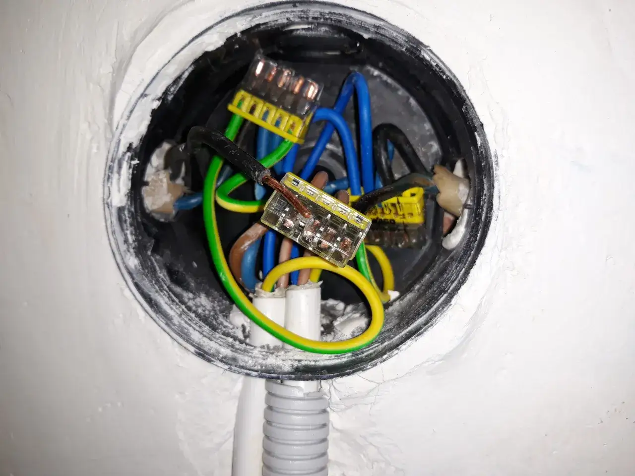 Widać przewody elektryczne w puszce, żółto-zielony, niebieski i czarny, połączone złączkami. To pomoc wizualna, jak podłączyć gniazdko w starej instalacji.